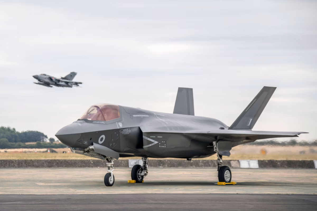 RAF F-35 Lightning II fighter jet parked on runway with another jet taking off in the background