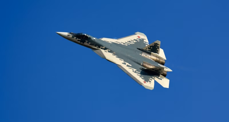 Sukhoi Su-57 aircraft number 054 in digital camouflage, photographed from below during flight against a clear blue sky