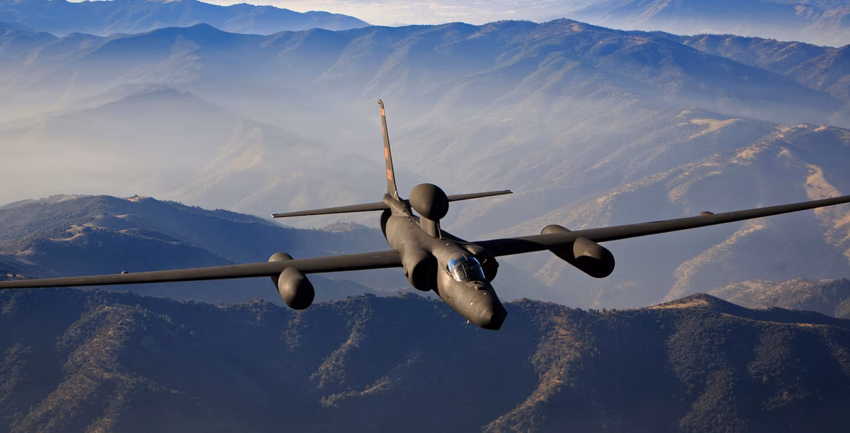 A U-2S Dragon Lady in flight over a mountain range, showing its distinctive glider-like wings and sensor pods.