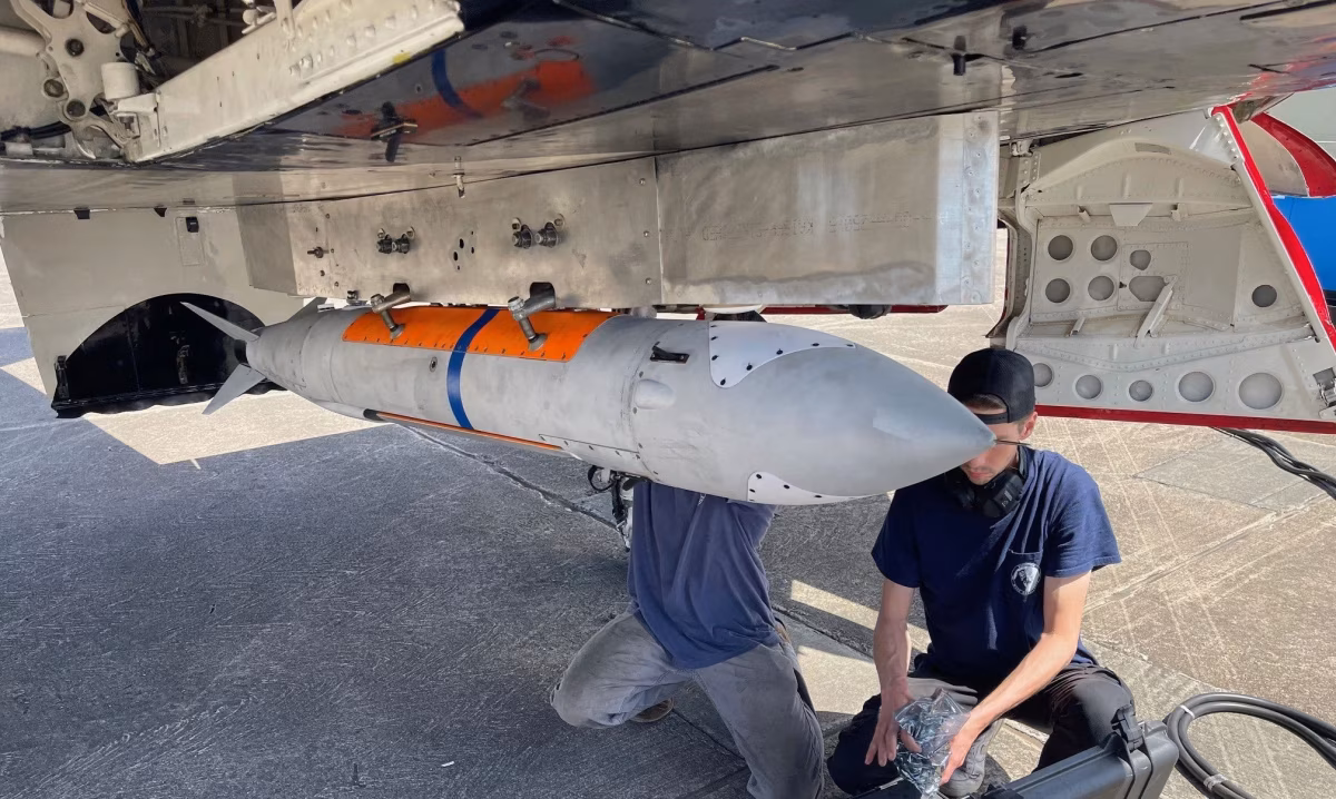 Two technicians inspecting a RAACM cruise missile mounted under the wing of an A-4 Skyhawk on the flight line, with an equipment case open on the ground beside them.