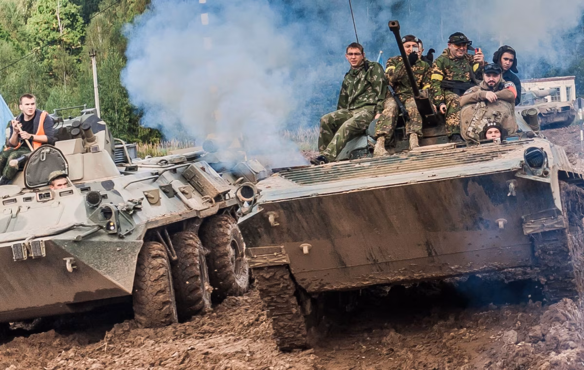 A wheeled BTR armored personnel carrier and a tracked BMP infantry fighting vehicle navigate muddy terrain side by side, with riders visible on top of both vehicles.