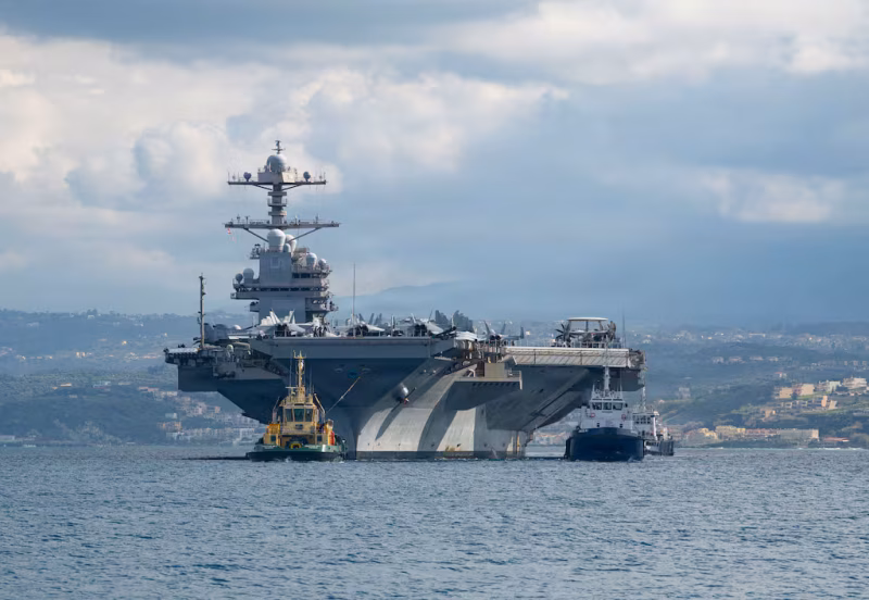 USS Gerald R. Ford aircraft carrier assisted by tugboats in Souda Bay, Greece, with coastal hills visible in the background