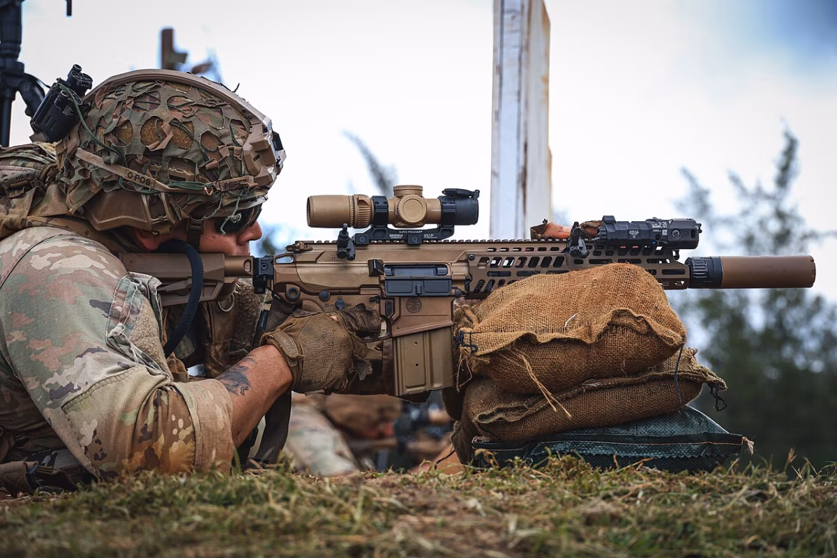 U.S. Army infantryman in full combat kit firing an M7 rifle from a prone position over sandbags, fitted with a Vortex AMG 1-10×24 LPVO scope, B.E. Meyers MAWL laser aiming module, and integrated suppressor, 2026