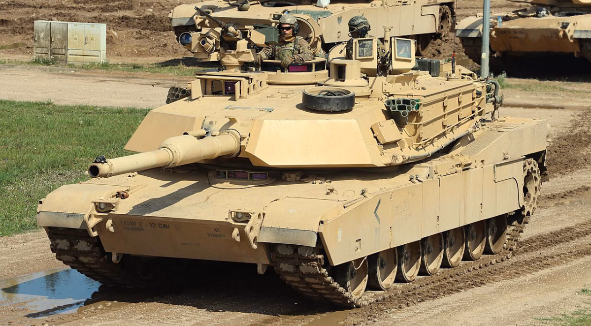 A U.S. Army M1A2 SEPv3 Abrams tank with crew visible in the commander's hatch, moving through muddy terrain during training operations in Poland, 2024. The CROWS remote weapon station and smoke grenade launchers are visible on the turret.