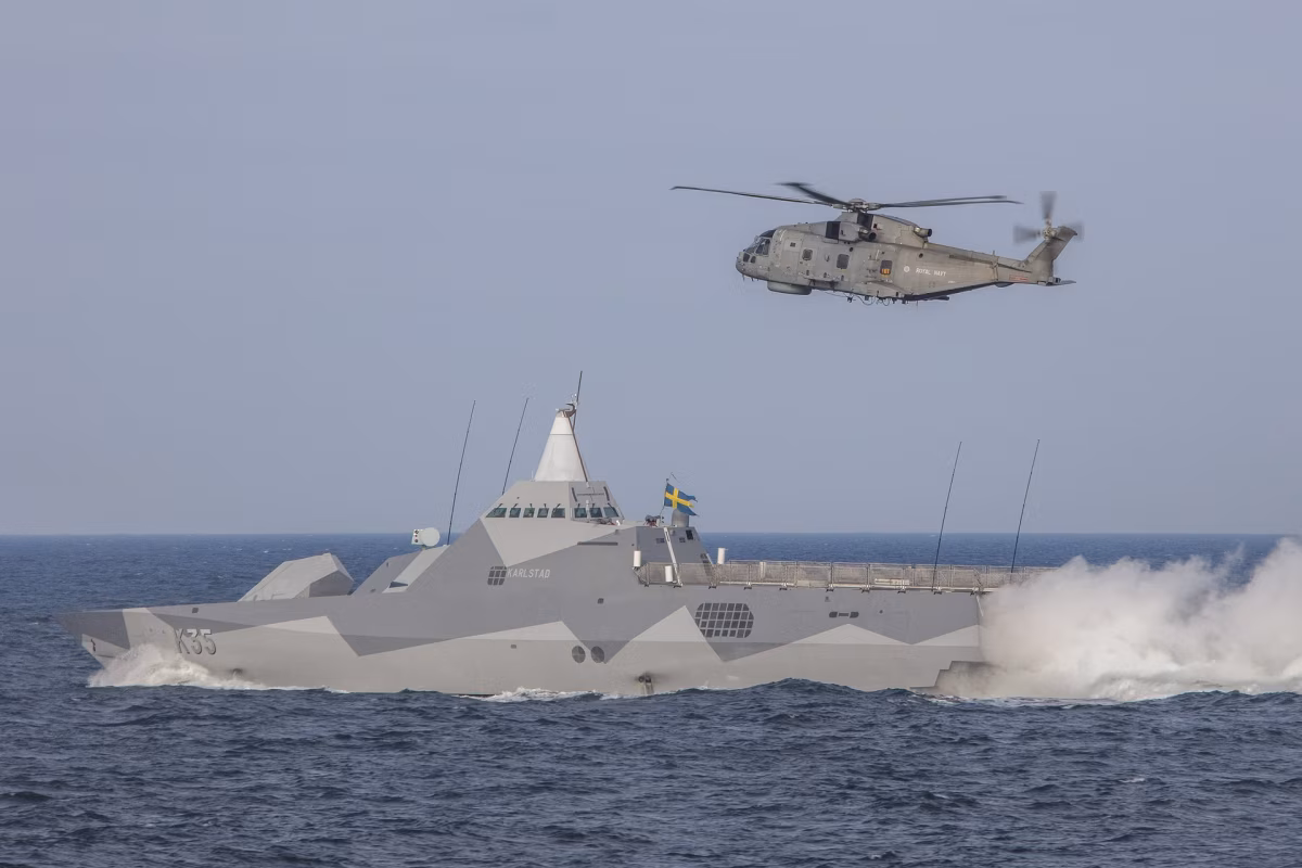 HSwMS Karlstad (K35) at speed during BALTOPS 2020, with a Royal Navy Merlin helicopter overhead