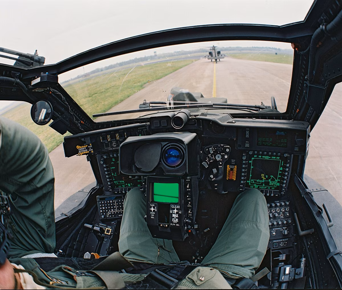 View from the gunner's seat inside an AH-64D Apache cockpit, showing the TADS monocle display, green-lit instrument panels, and a second Apache on the runway ahead.